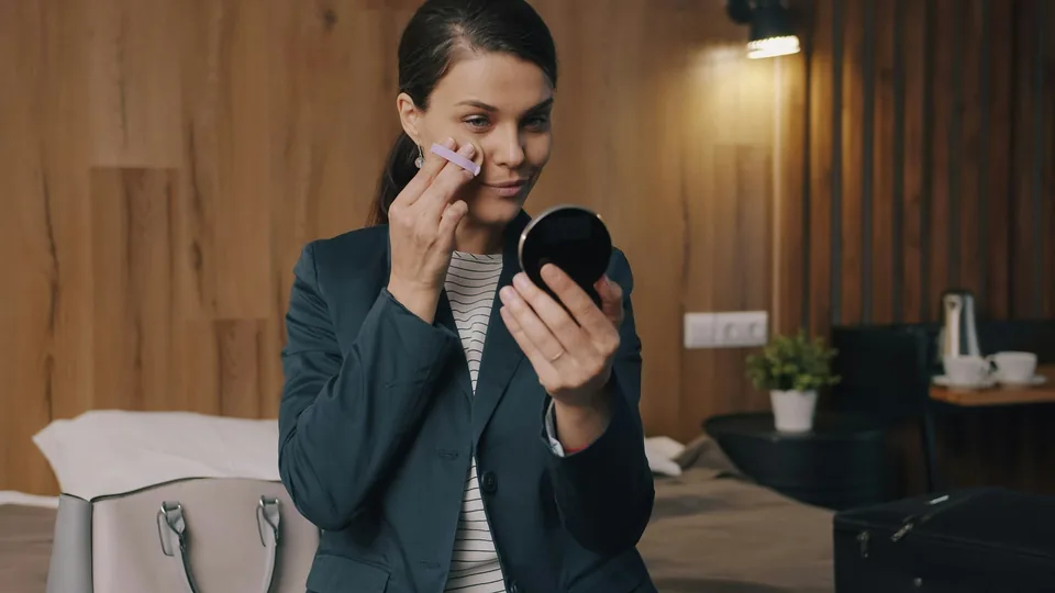 A woman in a dark blazer applying powder with a puff in a hotel room