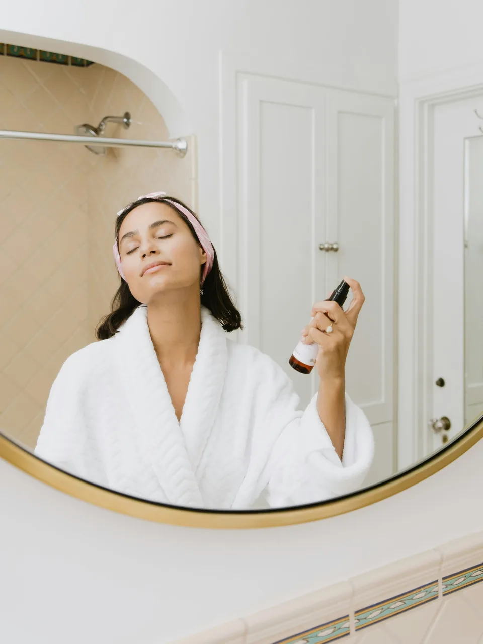 A woman in a white robe and headband spraying facial mist in a bathroom